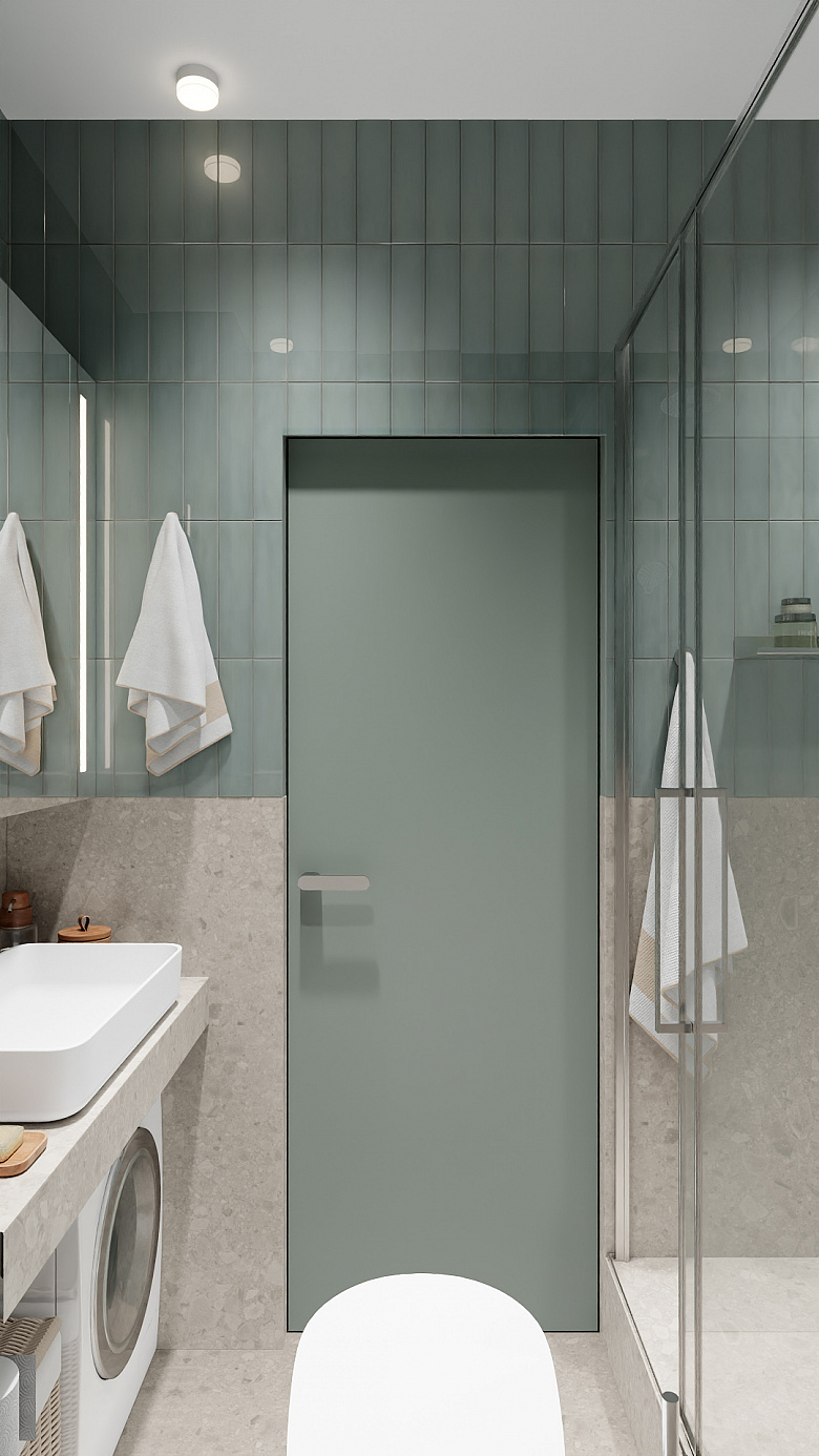 фото:Современная ванная в минималистичном стиле: приглушённая серо-зелёная плитка на стенах, светлая каменная фактура на нижней части стен и полу. Центральные элементы — стеклянная душевая кабина с хромированной фурнитурой, лаконичная прямоугольная раковина на подвесной столешнице, встроенный стиральный автомат. Акценты — аккуратно сложенные белые полотенца на хромированных держателях, матовая дверь в тон плитке. Точечное освещение и чистые линии создают ощущение простора и уюта. Функциональность без излишеств — ключевая идея дизайна