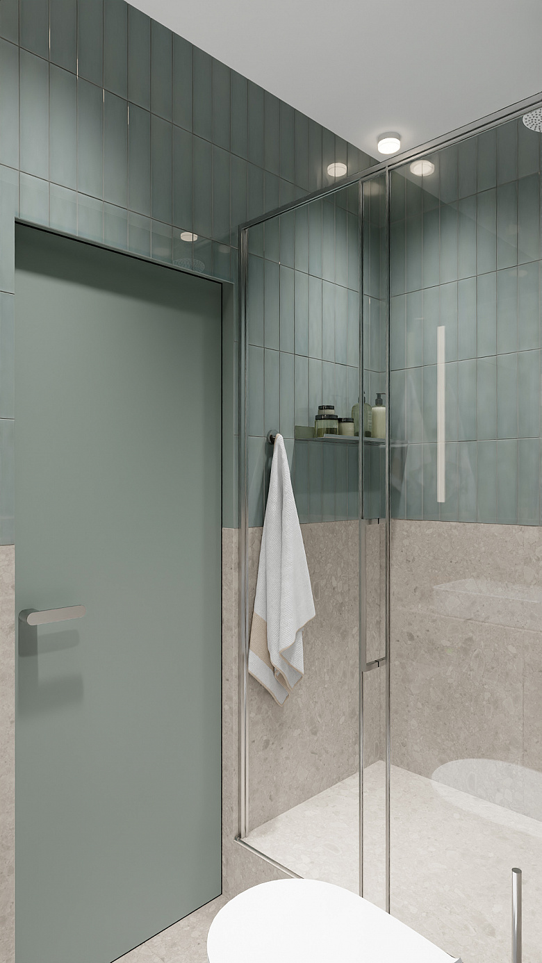 фото:Современная ванная в минималистичном стиле: сочетание матовой серо-зелёной плитки на стенах и светлого каменного покрытия в нижней части. Центральным элементом стала просторная душевая кабина с прозрачными стеклянными стенками и хромированным профилем. Акцент — лаконичная полка внутри душевой с аккуратно расставленными банными принадлежностями. Дополняют образ матовая дверь в тон плитке и белоснежное полотенце на стильном держателе. Мягкий рассеянный свет от встроенных светильников создаёт уютную атмосферу. Чистые линии и сдержанная палитра подчёркивают функциональность и элегантность пространства