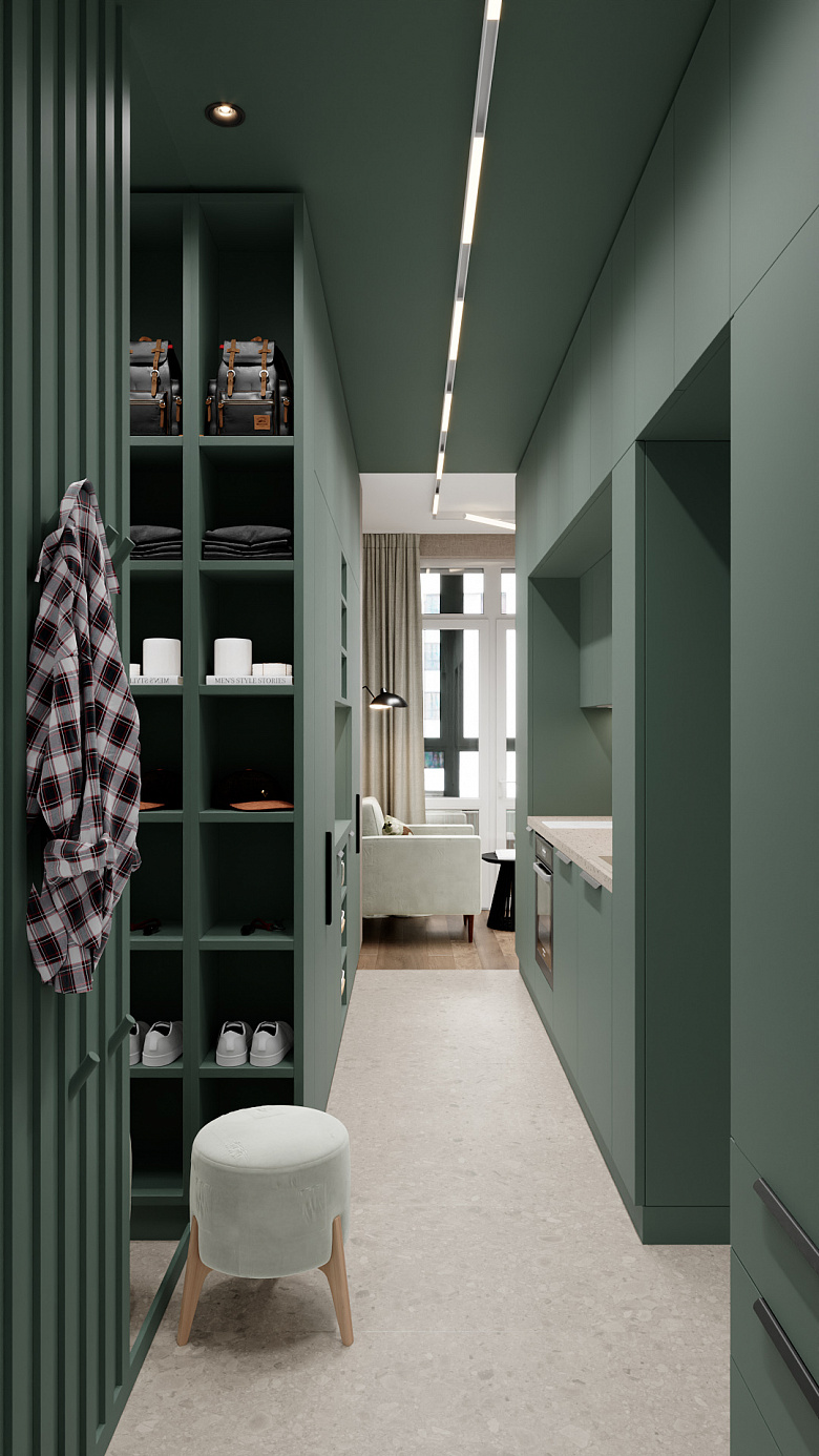 фото:Компактная прихожая в современном стиле с доминирующим глубоким зелёным цветом стен и мебели. Открытые стеллажи с аккуратно сложенными вещами и обувью, мягкий пуф на деревянных ножках для удобства. Линейное потолочное освещение подчёркивает геометрию пространства. Справа — фрагмент кухни в той же цветовой гамме, слева — оригинальная вешалка. Чистые линии, функциональность, сдержанная цветовая палитра создают ощущение порядка и уюта