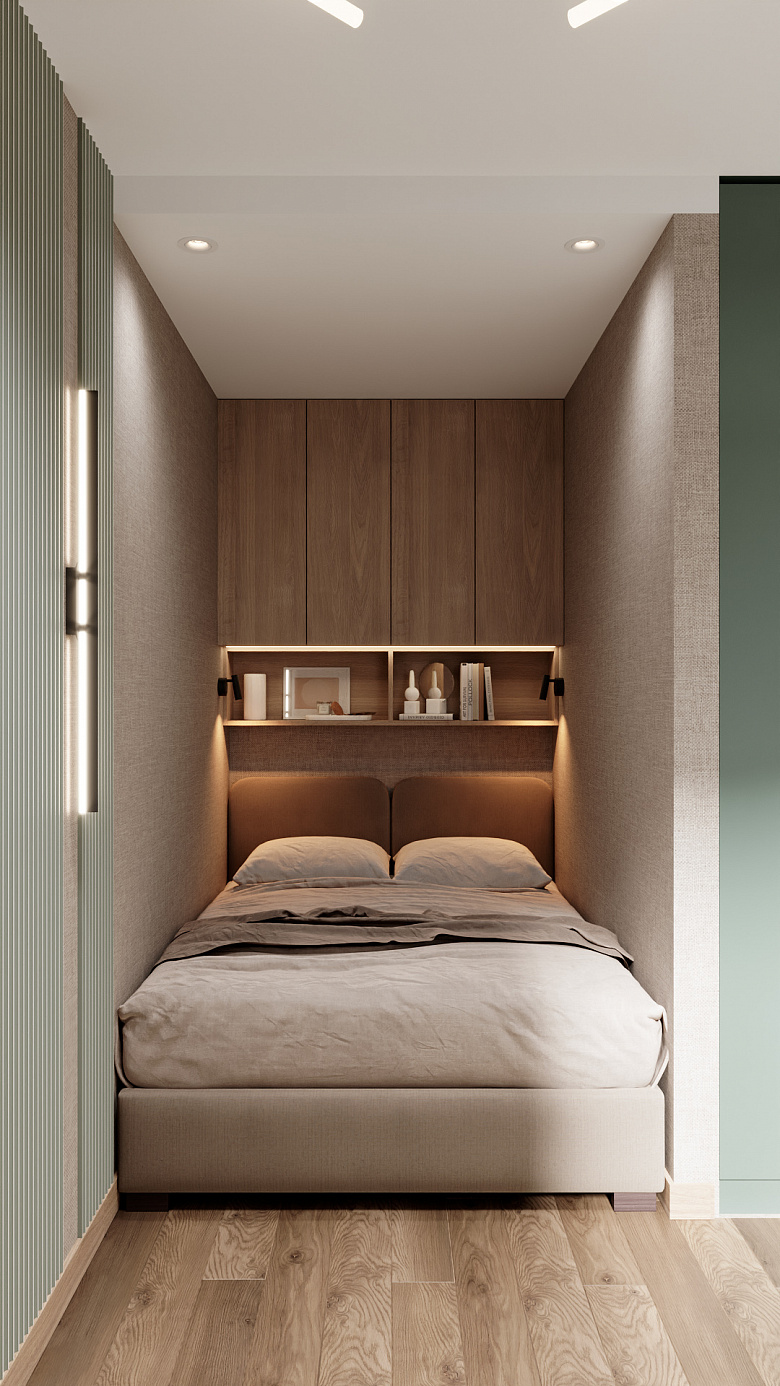 фото:Компактная спальня в минималистичном стиле: кровать с мягким изголовьем встроена в нишу, над ней — навесной шкаф и открытая полка с подсветкой для декора и книг. Стены в нейтральных бежево-серых тонах, акцент — вертикальные рифлёные панели в приглушённом зелёном оттенке. Пол — светлый паркет, мягкое освещение создаёт уютную атмосферу. Лаконично, функционально, уютно
